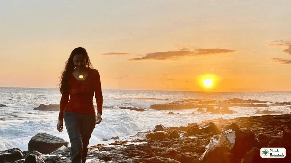 Woman walking along a rocky shoreline at sunset with a small glowing light at her chest