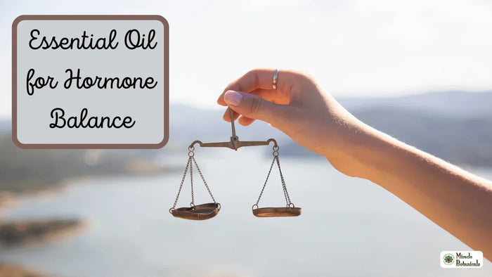 Hand holding a small brass balance scale against a blurred lake view, with text: Essential Oil for Hormone Balance